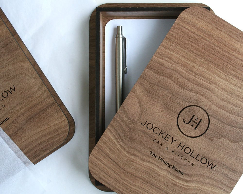 A wooden check presenter box with a pen, opened to show the interior which has a slot for a tip and a compartment for a business card or logo.