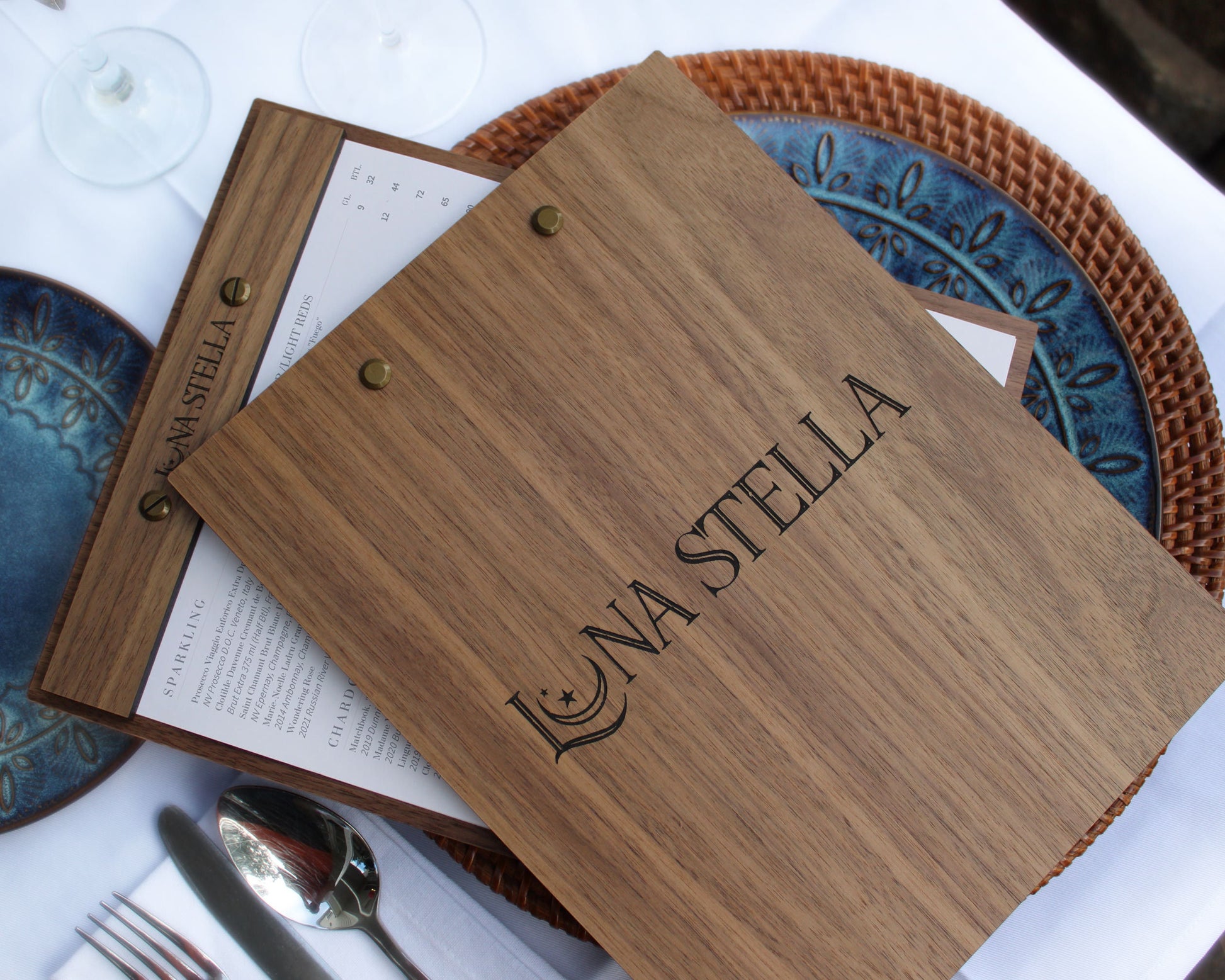 A walnut wood menu holder with a customized engraved logo on the front, displayed on a table with a menu and a basket in the background.